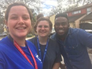 Three people from the Florida Field Team smile outdoors—two women and one man. The woman on the left wears blue, the middle woman has glasses, and the man on the right wears a denim shirt with braces. Houses and trees fill the background.
