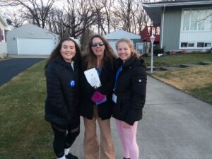 Three women from the Indiana Field Team stand together and smile outdoors in a suburban neighborhood. Two wear black jackets and name tags, while the woman in the middle wears sunglasses and holds a pamphlet. Bare trees and houses are in the background.