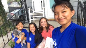 Four smiling young women from the Indiana Field Team sit on a porch, three in matching blue shirts, one in pink, as one holds a small gray dog. They appear happy, with one taking a selfie and holding papers by the house entry steps.