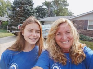 Two women from the Ohio Field Team, wearing blue shirts, smile outside in a suburban neighborhood with houses, trees, and cars visible in the background.