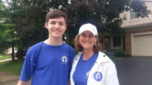 A young man and a woman from the Ohio Field Team smile outdoors, both wearing matching blue shirts with a white logo and text. The woman also wears a white jacket and hat. They stand on a suburban street in front of a house with trees.