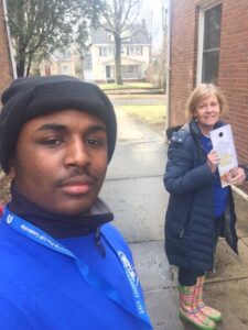 A young man takes a selfie outdoors with a woman in a coat and colorful rain boots, holding an Ohio Field Team flyer. They stand on a wet sidewalk near brick buildings and houses in the background.