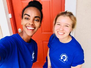 Two smiling women in matching blue Florida Field Team t-shirts stand in front of a red door, taking a selfie together. The shirts have a white logo on the left side. Both women appear happy and are looking at the camera.
