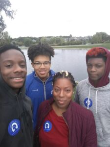 Four people from the Indiana Field Team stand close together, smiling by a calm lake with houses and trees in the background. All wear jackets or hoodies and blue awareness stickers. The sky is overcast.