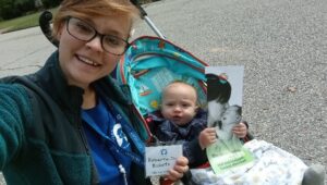 A smiling woman with glasses, wearing an Indiana Field Team name badge that reads “Roberta-Joy Ricketts,” takes a selfie next to a baby in a stroller. The baby holds a flyer as they enjoy time outdoors on a paved surface.
