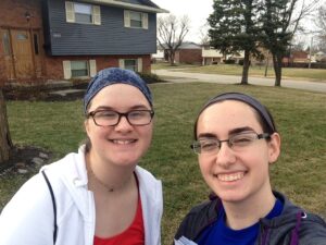 Two people from the Ohio Field Team smile outdoors in front of a suburban house with a yard and tree. Both wear glasses and headbands, enjoying the clear sky.