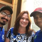 Three people smile at the camera near a building entrance in West Virginia. Two men from the Field Team wear blue shirts and hats, while the woman in the center wears a blue and white patterned shirt.