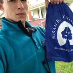 A young person in a turquoise fleece, part of the Field Team, holds up a blue drawstring bag that reads “Susan B. Thun— Elections & Law,” standing outside a house in West Virginia.