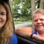 Two women smiling; one sits inside a car while the other—part of the Field Team—stands outside next to the window, with trees and a suburban West Virginia street in the background. It appears to be a friendly outdoor encounter.