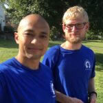 Two men wearing matching blue T-shirts stand outside on a sunny day in West Virginia, smiling at the camera. They are part of a Field Team on a grassy lawn, with trees and houses visible in the background.