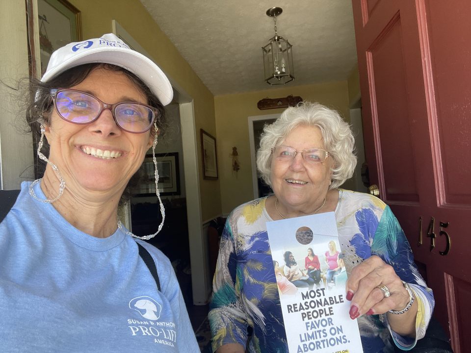 Two smiling women pose at a front door. One wears a Susan B. Anthony Pro-Life America shirt and cap; the other holds a pamphlet reading Most Reasonable People Favor Limits on Abortions.