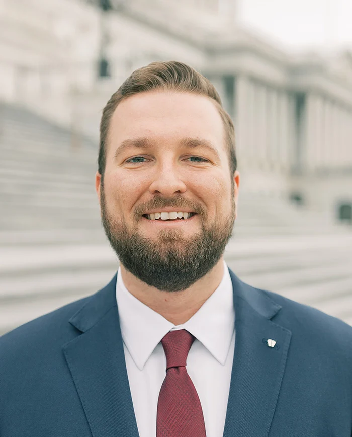 A man with short brown hair and a beard, wearing a blue suit, white shirt, and red tie, smiles while standing outside in front of stone steps and a large building.