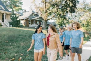 A group of young adults and a woman, possibly part of a Title X or Planned Parenthood initiative, walk and laugh together on a sunny residential sidewalk in matching light blue T-shirts. Houses and trees are visible in the background.