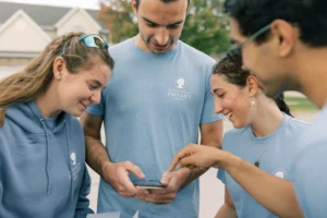 Four young adults wearing blue “Pro-Life” shirts smile and look at a smartphone outdoors, as one points at the screen. Houses and green trees are visible in the background, possibly discussing taxpayer dollars or Planned Parenthood.