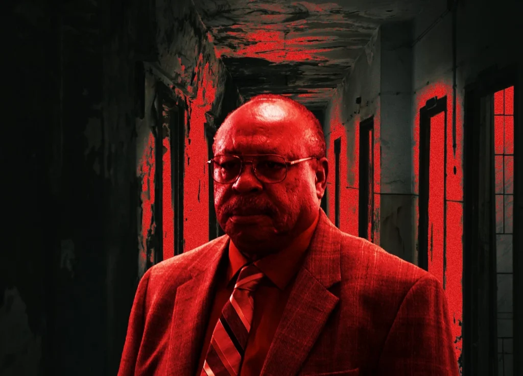 An older man wearing glasses, a suit, and a striped tie stands in a dark, decrepit hallway with red lighting, reminiscent of the intense media coverage surrounding convicted murderer Kermit Gosnell.