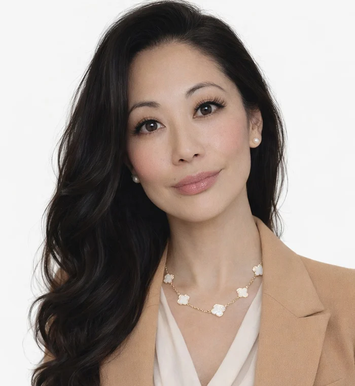 Kelsey Pritchard, with long dark hair, wears a beige blazer, cream blouse, pearl earrings, and a delicate gold necklace as she poses against a white background, smiling gently at the camera.