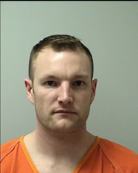 A man with short light brown hair, wearing an orange prison uniform, is facing the camera against a plain light-colored background.