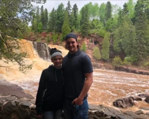 A couple stands in front of a rocky river and waterfall, surrounded by lush green trees and foliage. The sky is cloudy, and both people are smiling at the camera.