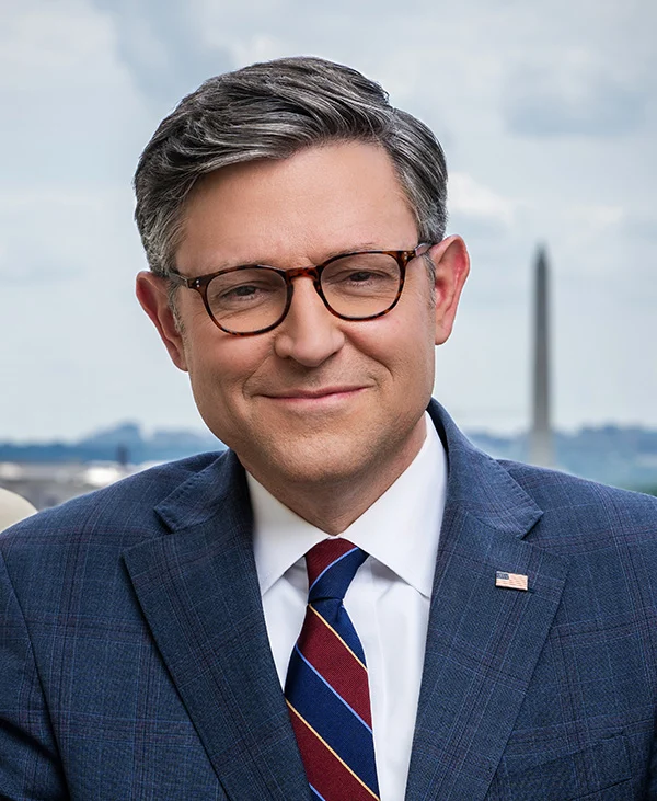 A man with short gray hair and glasses, wearing a blue suit, white shirt, and striped tie, smiles in front of a blurred cityscape with the Washington Monument in the background, reminiscent of a classic portrait of Mike Johnson.