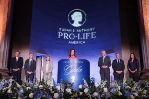 A woman speaks at a podium decorated with flowers at the Susan B. Anthony Pro-Life America Gala, with Dannenfelser and five others standing behind her on stage and the event logo projected on the wall.