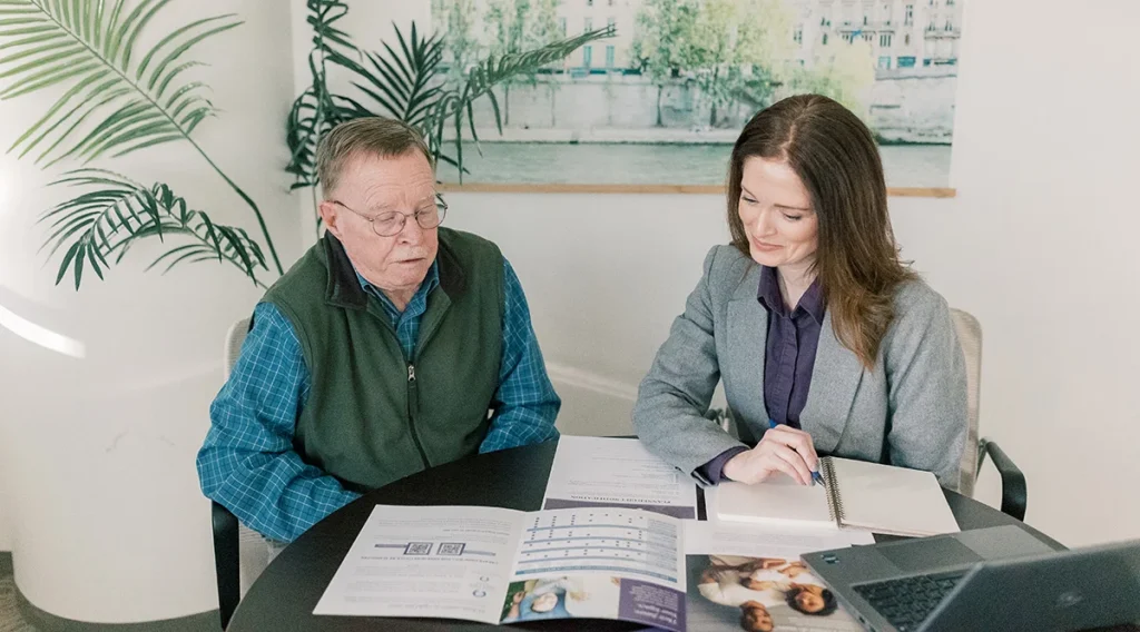 A woman in business attire sits at a table with an older man, reviewing documents together. Papers and a laptop are on the table. They appear to be discussing information in a bright, modern office setting.
