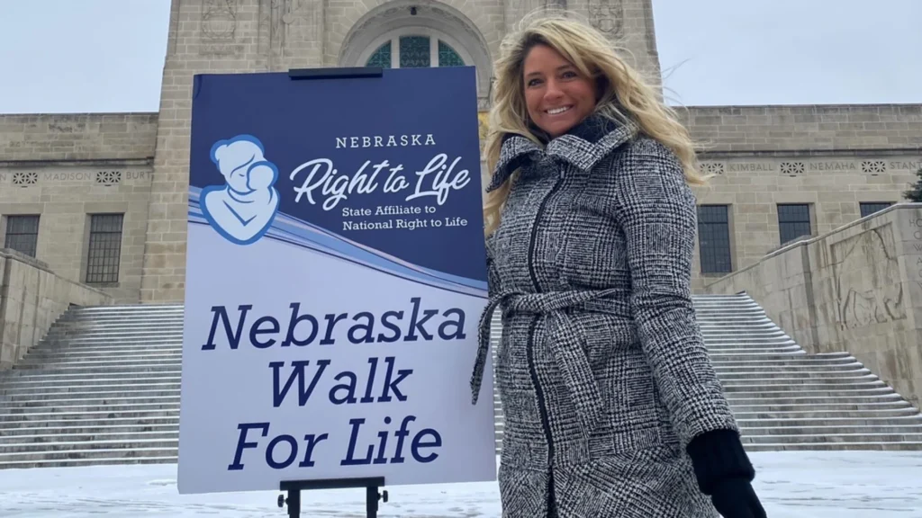 A smiling woman, Haile, in a gray coat stands next to a Nebraska Walk For Life sign outside a large stone building on a snowy day, sharing her survival story about the dangers of abortion drugs.