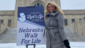 A smiling woman, Haile, in a gray coat stands next to a Nebraska Walk For Life sign outside a large stone building on a snowy day, sharing her survival story about the dangers of abortion drugs.