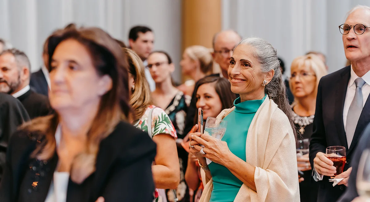 A group of well-dressed people at an indoor event. A smiling woman in a turquoise dress and white shawl stands in the center, holding a drink, while others around her watch attentively, celebrating her inspiring pro-life legacy.