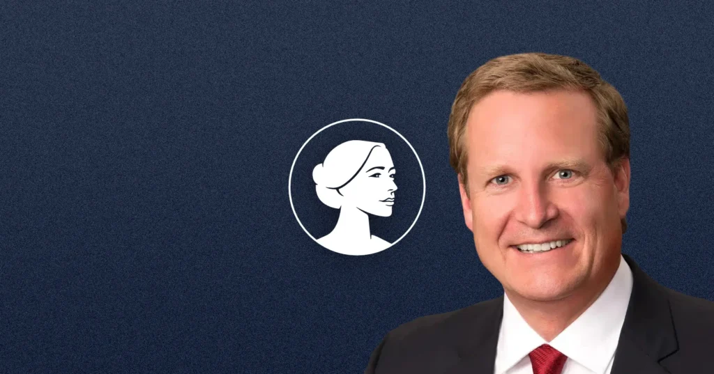 A smiling man in a suit and red tie stands before a dark blue background with a white stylized woman’s profile logo next to him, representing MT-Sen candidate Kurt Alme and his strong Pro-Life stance.