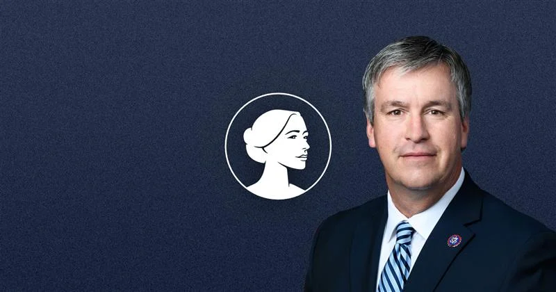 A man in a suit and striped tie stands in front of a dark blue background with a white profile illustration of a woman's face in a circle to his left, representing Barry Moore and the Pro-Life message in the AL-Sen Race.