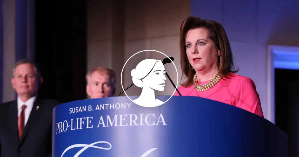 A woman in a pink dress speaks at a podium labeled Susan B. Anthony Pro-Life America, as Dannenfelser and another man stand in the background. A logo featuring a woman’s profile is overlaid, emphasizing the debate on abortion policy.