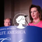 A woman in a pink dress speaks at a podium labeled Susan B. Anthony Pro-Life America, as Dannenfelser and another man stand in the background. A logo featuring a woman’s profile is overlaid, emphasizing the debate on abortion policy.