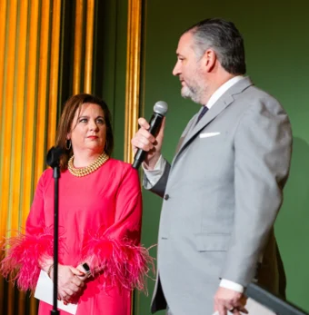A woman in a pink dress with feathered sleeves stands next to a man in a light gray suit, speaking into a microphone at the SBA Gala, set against a striking green and gold backdrop.