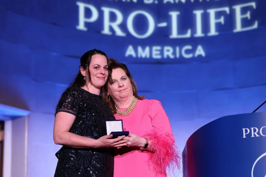 Two women stand together on stage at the SBA Gala, one holding an award box. Both are dressed formally, smiling for the camera. The backdrop reads PRO-LIFE AMERICA in large letters, celebrating life and achievement.