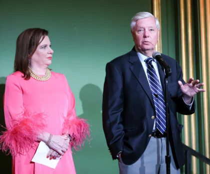A woman in a pink dress with feathered sleeves stands beside a man in a dark blazer and striped tie speaking into a microphone at the SBA Gala; they are in front of a green wall.