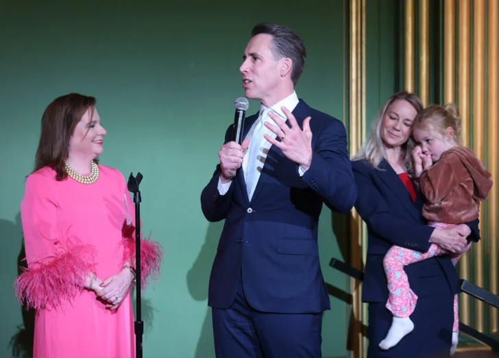 A man in a suit speaks into a microphone while gesturing, standing next to a woman in a bright pink dress at the SBA Gala. Nearby, another woman holds a young child in her arms, capturing the warmth of life and family meeting moments against a green and gold wall.