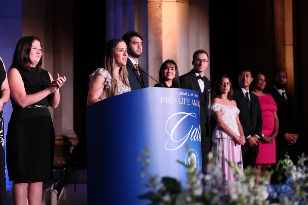 A woman speaks at a blue podium labeled Pro-Life Awards Gala during the SBA Gala, while several men and women stand behind her on stage, some applauding, in a formal setting with columns and evening attire.