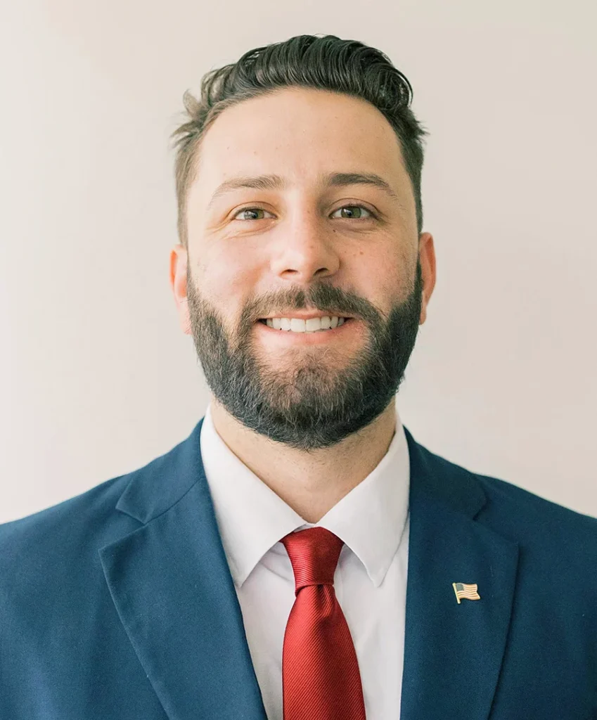 Parker Dillon, a smiling man with a beard, wears a blue suit, white shirt, and red tie with a flag pin on his lapel as he stands against a plain light background.