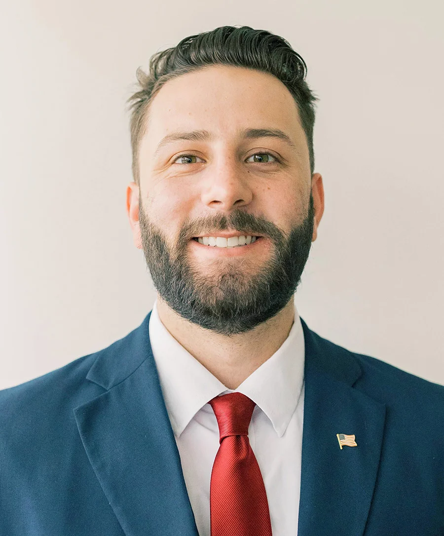 Parker Dillon, a smiling man with a beard, wears a blue suit, white shirt, and red tie with a flag pin on his lapel as he stands against a plain light background.