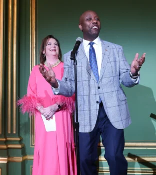 At the SBA Gala, a man in a gray plaid suit jacket speaks into a microphone, gesturing with his hands, while a woman in a bright pink dress with feathered sleeves stands behind him, smiling. The scene captures the vibrancy of life in an elegant, ornately decorated room.