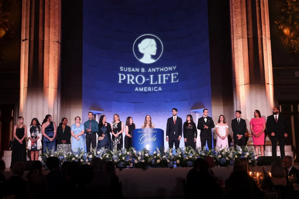 A group of people stands on stage behind a podium with the sign SBA Gala at a formal event. The backdrop displays the Susan B. Anthony Pro-Life America logo, celebrating Life, with the stage decorated with white and blue flowers.
