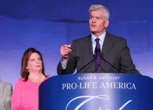 A man in a gray suit speaks at a podium labeled Susan B. Anthony Pro-Life America while a woman in a pink top stands nearby, looking at him. The background is blue and purple.