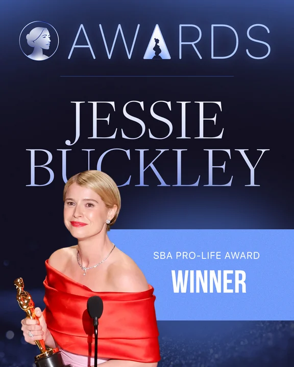 A woman in a red off-the-shoulder gown holds a gold award statue, standing before a sign that reads AWARDS Jessie Buckley SBA Life Awards Pro-Life Award Winner.