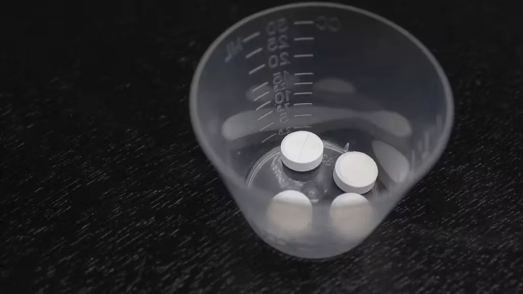A close-up of a small, clear plastic medicine cup containing three round white tablets on a dark surface, hinting at how someone may have sought abortion drugs online.
