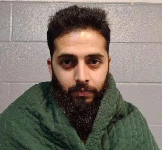 A man with a full beard and short hair sits wrapped in a green blanket against a gray concrete block wall, looking directly at the camera with a neutral expression.