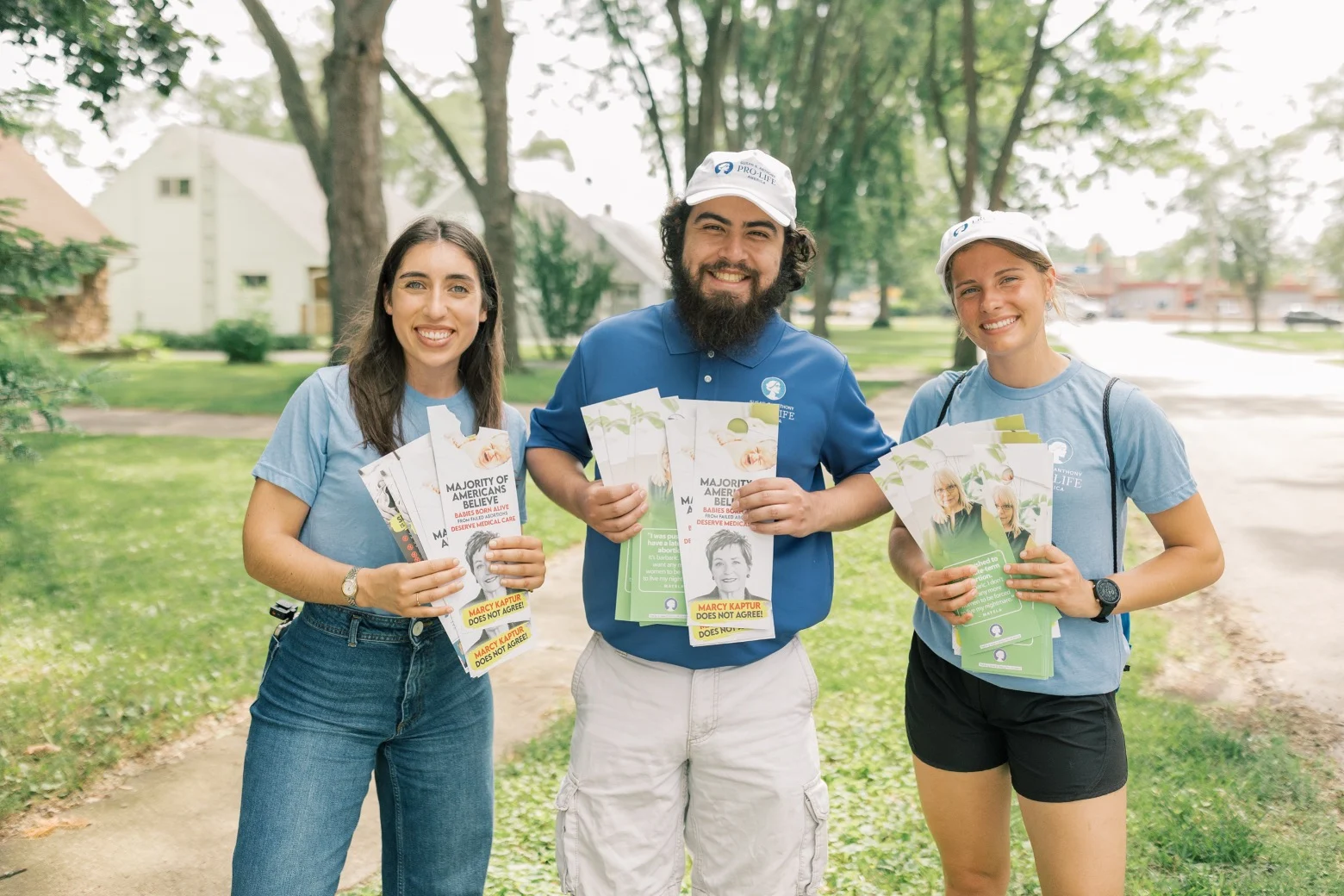 Three smiling people stand outdoors in Toledo, Ohio, holding campaign flyers and wearing blue shirts while canvassing for SBA Pro-Life America. The man in the center wears a hat, as the two women beside him look at the camera with trees and houses behind them.