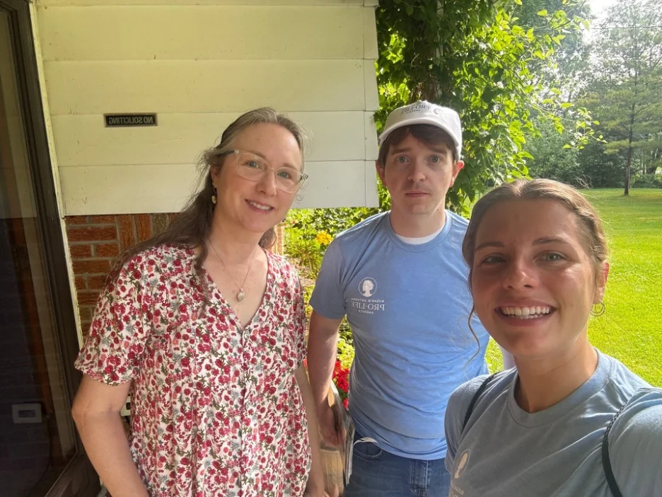 Three people pose and smile outside near a house in Toledo, Ohio. One woman wears glasses and a floral dress, a man in a blue shirt and cap stands in the middle, and another woman in a blue tee smiles in the foreground. A green lawn is visible behind them.