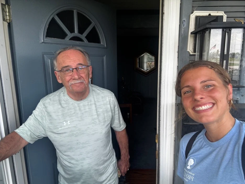 A smiling young woman takes a selfie with an older man standing in the doorway of a house in Toledo, Ohio. The man is wearing glasses and a light-colored shirt, while the woman, dressed in blue, looks happy after canvassing.