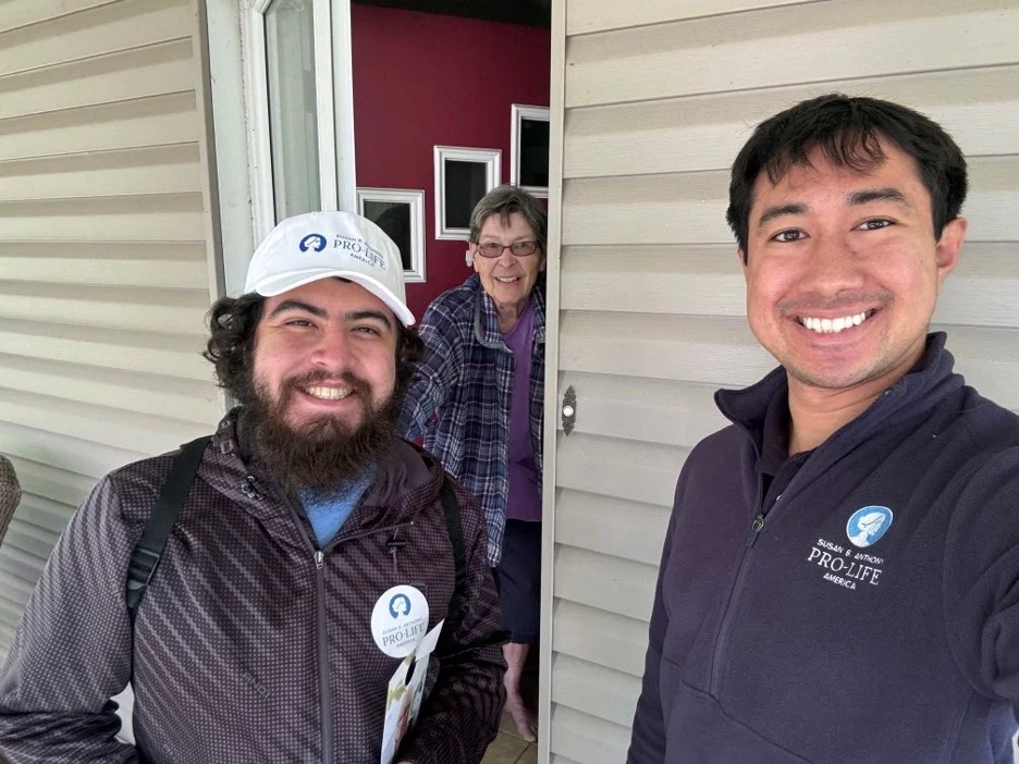 Two smiling men with “Pro-Life” badges, canvassing for SBA Pro-Life America, stand outside a house in Toledo, Ohio; one takes a selfie as the other holds papers. An older woman, also smiling, appears in the doorway behind them.