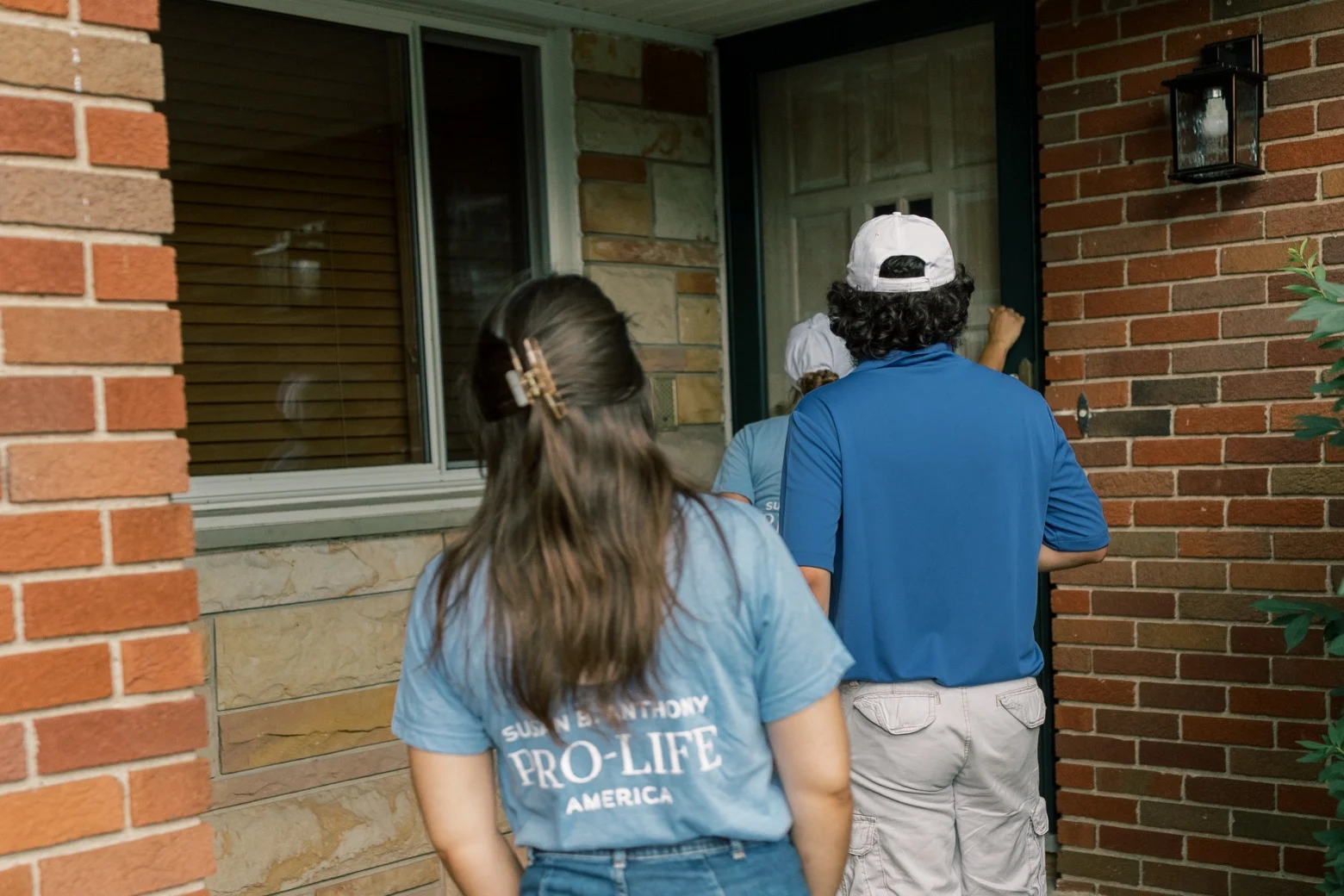 Three people stand at a brick house entrance in Toledo, Ohio; one knocks on the door. The person in front wears a blue shirt and white cap, while another’s shirt reads SBA Pro-Life America, suggesting they are canvassing the neighborhood.